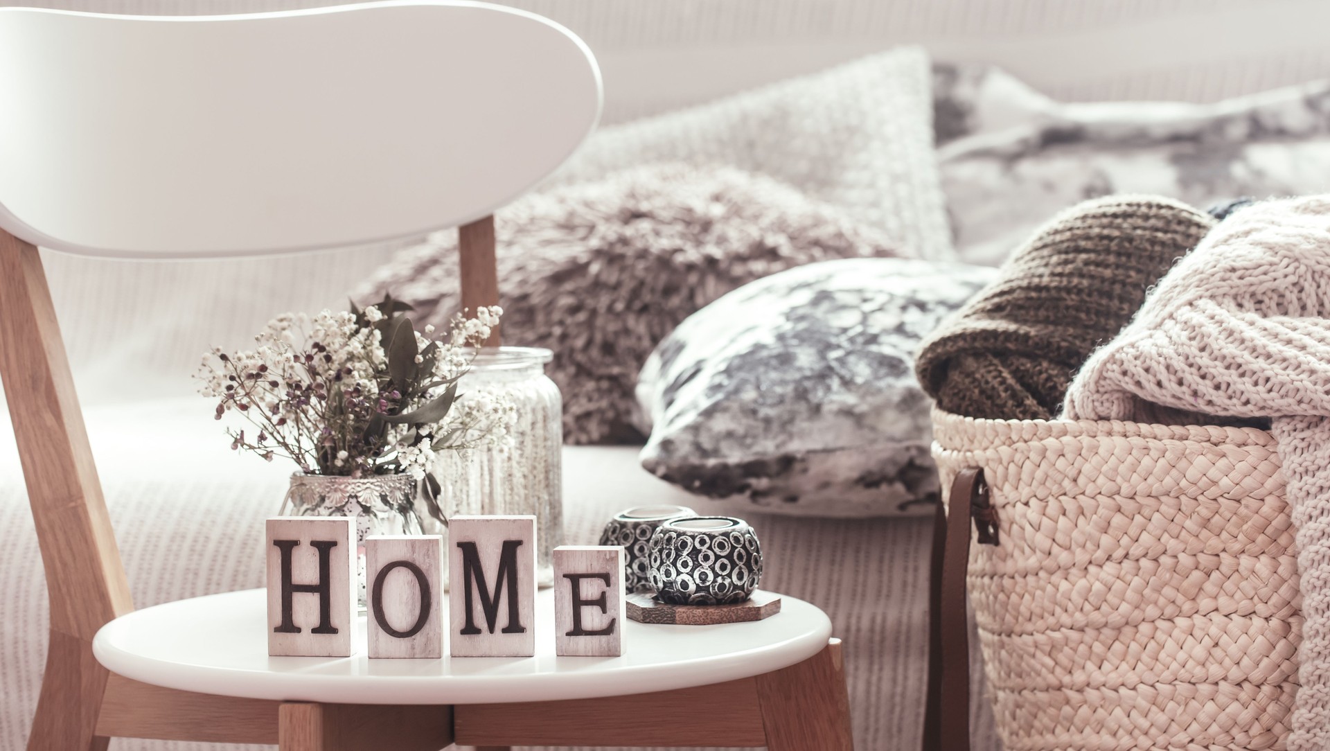 Candles, a vase with flowers with wooden letters of the home on wooden white chair. Sofa and wicker basket with cushions in the background.